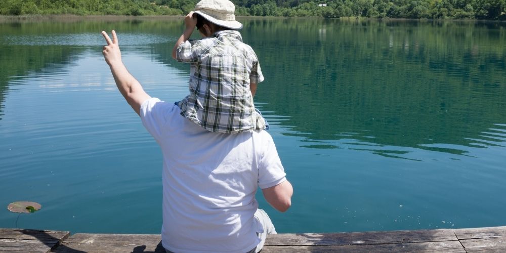 A young boy sitting on his dad's shoulder on a dock, looking out onto a lake.