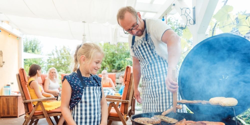 Dad grilling with his daughter watching and the rest of his family sitting at a table in the background.
