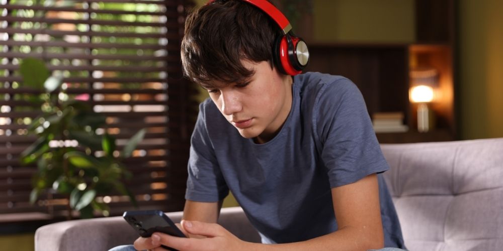 Teen boy sitting on the couch, wearing headphones while scrolling on his phone.