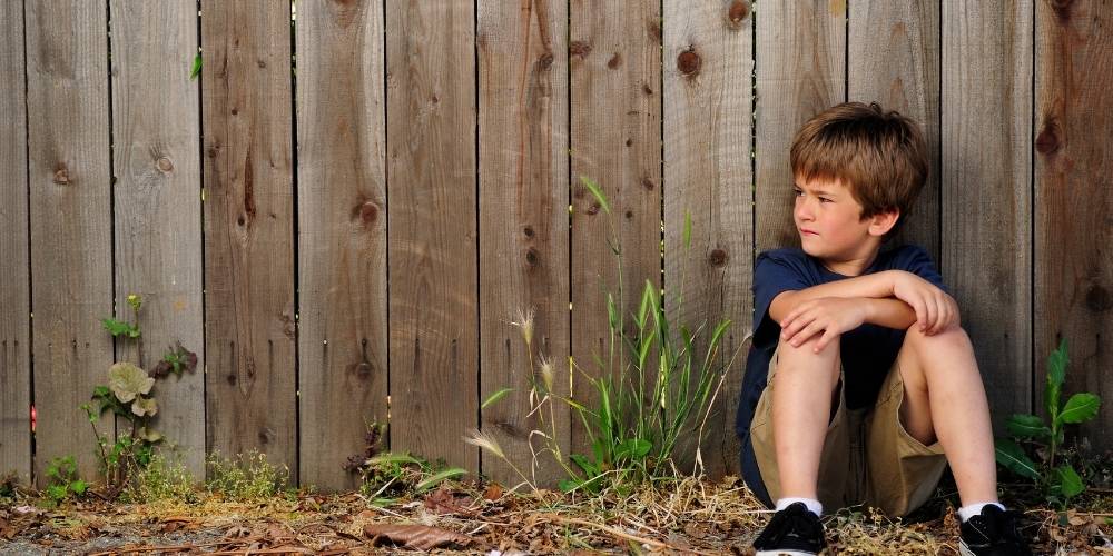 A young boy sitting outside against a fence alone.