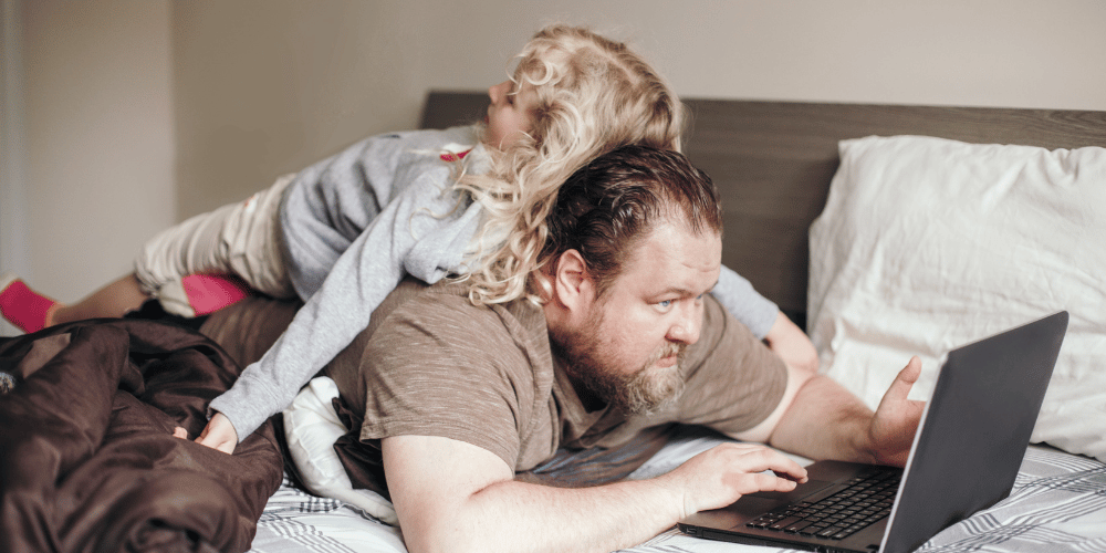 A dad is lying stomach down on his bed working on his laptop while his young daughter is lying on his back.